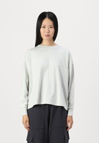 Světle šedá dlouhá košile s kulatým výstřihem, volný střih a spadlé ramena, v kombinaci s tmavými cargo kalhotami. Hladká textura látky.