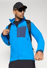 Giacca in fleece blu con tasca sul petto con zip, tessuto testurizzato e colletto alto, indossata sopra una maglietta nera con uno zaino.
