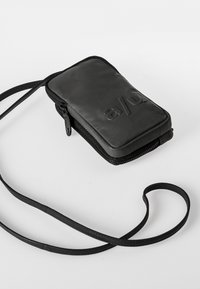 Schwarze Reißverschluss-Tasche aus glattem Material, mit verstellbarem Riemen und geprägtem Logo auf der Vorderseite, kompakte rechteckige Form.
