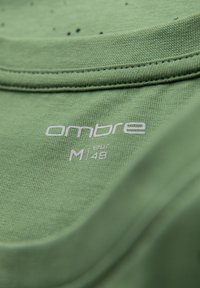 Tela de camiseta de algodón verde con una textura suave. Presenta la marca "ombre" y la talla "M" impresas en blanco en el cuello.