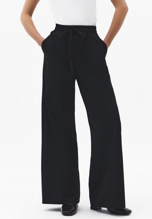 Personne portant un pantalon large noir ample avec une taille élastique et un cordon de serrage, les mains dans les poches, associé à des chaussures noires.