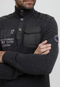 Pilkos megztos palaidinės su aukštu apykaklės, užsegimu mygtukais ir pečių detalėmis. Yra krūtinės kišenė ir siuvinėtas logotipas.