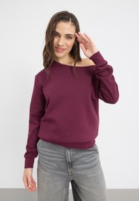 Burgundska dukserica bez ramena s mekim teksturom, dugim rukavima i opuštenim krojem, u kombinaciji s rastegnutim sivim trapericama. Minimalistički dizajn.