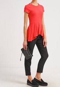 Röd peplumtop med korta ärmar, svarta slim-fit byxor och svarta plattformssneakers, håller en svart clutchtask.