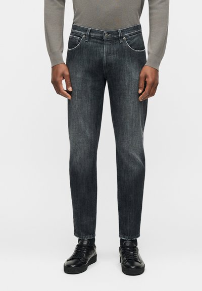 Άνδρας που φοράει ξεθωριασμένο μαύρο τζιν με ίσια γραμμή, γκρι πουκάμισο με μακριά μανίκια και μαύρα δερμάτινα αθλητικά παπούτσια, στέκεται μπροστά σε λευκό φόντο.