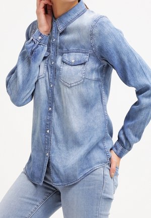 Vrouw draagt een lichtblauw denim overhemd met borstzakken en bijpassende jeans, poseert met één hand nabij de nek en de andere in de zak.