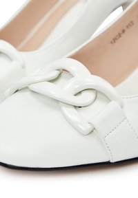 Chaussures plates en cuir blanc avec un grand détail en maillon de chaîne brillant sur le devant et un design d'orteil carré arrondi.