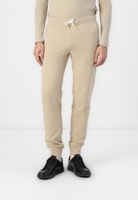 Beige joggebukser med snor i livet, tettsittende mansjetter og en myk tekstur, kombinert med svarte sneakers.
