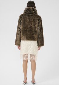 Barna műszőrme kabát magas gallérral, áttetsző, hímzett krém színű ruhával és ezüst magas sarkú cipőkkel párosítva.
