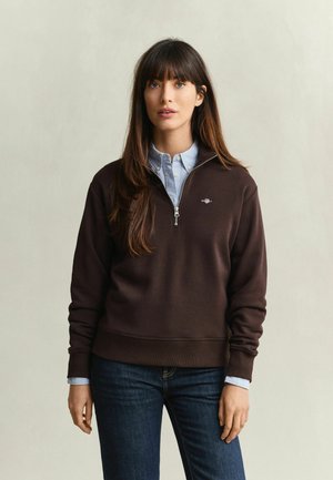 Femme aux longs cheveux bruns portant un sweat-shirt marron foncé à demi-fermeture éclair sur une chemise bleu clair à col et un jean foncé, debout devant un fond neutre.