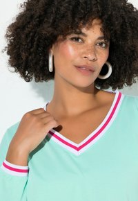 Donna con capelli ricci che indossa grandi orecchini a cerchio e una maglietta verde menta con scollo a V a righe bianche e rosa e maniche a sbuffo.