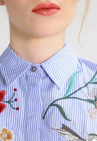 Niebieska i biała koszula w paski z wykładanym kołnierzykiem, z haftem kwiatowym w kolorze czerwonym, zielonym i żółtym oraz pojedynczym zapięciem na guzik.