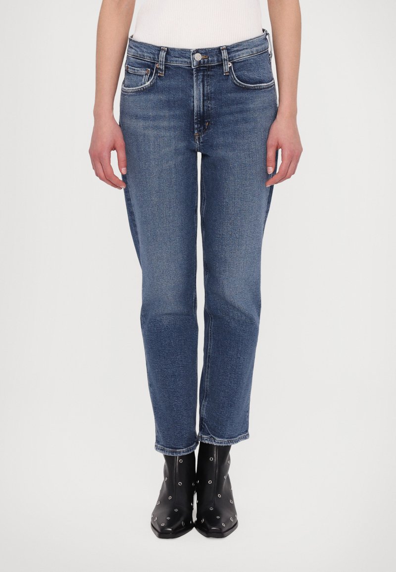 Tumedat sinised denim-jopeid sirge jalaga ja kergelt tekstureeritud. Omavad esitaskuid ja nööbikinnitust, kantud mustade pahkluude saapadega.