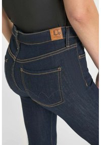 Diepblauwe hoge taille jeans met oranje stiksels, voorzien van een leren patch in bruin, achterzakken en een subtiel decoratief patroon op de zak.