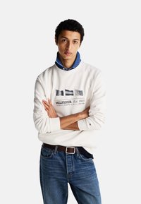 Sudadera blanca de manga larga con un gráfico en tonos azul marino y gris, combinada con jeans de mezclilla azules y una camisa de mezclilla con cuello debajo.