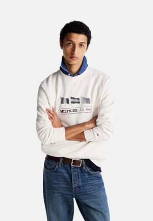 Sudadera blanca de manga larga con un gráfico en tonos azul marino y gris, combinada con jeans de mezclilla azules y una camisa de mezclilla con cuello debajo.