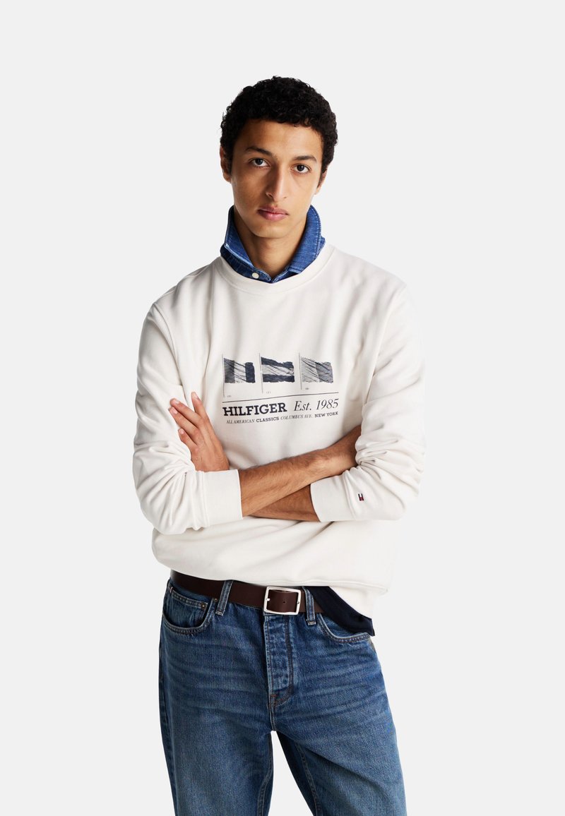 Sudadera blanca de manga larga con un gráfico en tonos azul marino y gris, combinada con jeans de mezclilla azules y una camisa de mezclilla con cuello debajo.