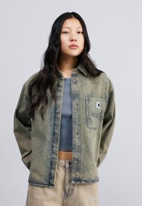 Denim jacket with a faded olive green color, oversized design, and a chest pocket featuring a small logo. Styled with a ribbed gray top.