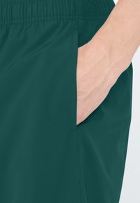 Vihreät urheilushortsit, joissa on joustava vyötärö ja sivutaskut, valmistettu sileästä ja kevyestä kankaasta, jossa on hienostunut kiilto.