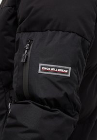 Černá péřová bunda s prošívanou texturou. Obsahuje kapsu na zip a logo nášivku s nápisem "KINGS WILL DREAM" na rukávu.