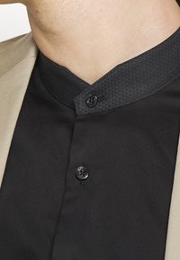 Camicia nera con colletto alto e texturizzato. Presenta due bottoni neri e una giacca beige di lato. Tessuto liscio con un design elegante.
