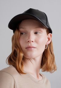 Căciulă din piele neagră cu o margine curbată, având detalii de cusătură. Un model cu părul castaniu, ondulat, până la umeri o poartă. Design simplu, minimalist.