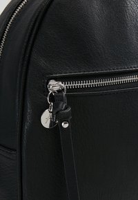 Sac à dos en cuir noir avec une fermeture éclair argentée, un curseur rond et une petite étiquette métallique à logo attachée à la fermeture éclair. Texture lisse.