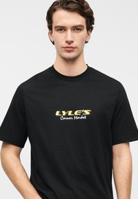 Μαύρο βαμβακερό T-shirt με γραφικό μπροστά που γράφει "LYLE'S" σε χρυσό και "Corner Market" σε λευκό, λαιμόκοψη με ρεβέρ, άνετη εφαρμογή.