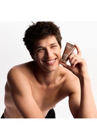 Een bruine tube van een NUXE huidverzorgingsproduct voor mannen, vastgehouden door een lachende persoon. De achtergrond is wit. De tube heeft een eenvoudig ontwerp en duidelijke etikettering.