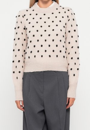 Femme portant un pull tricoté rose pâle à pois noirs et un pantalon ample taille haute gris foncé.