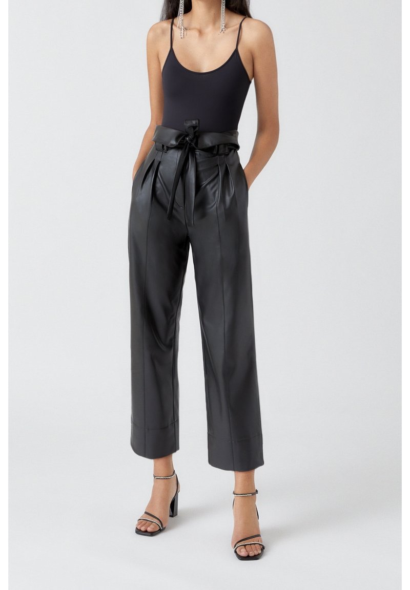 Femme portant un haut noir sans manches, un pantalon en cuir noir taille haute style paperbag avec ceinture, et des talons blocs noirs ouverts à l’avant avec brides à la cheville.