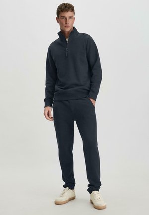Marineblauwe rits sweatshirts en bijpassende joggingbroek. Katoenmengsel, geribbelde manchetten. Model draagt een crème sneakers, handen in de broekzakken.