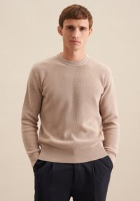 Beige gerippter Strickpullover mit Rundhalsausschnitt und langen Ärmeln, kombiniert mit dunklen Hosen. Weicher Stoff und dehnbares Material sind sichtbar.