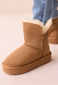 Stivaletto alla caviglia in pelle scamosciata marrone con un morbido rivestimento interno in pelliccia sintetica crema, caratterizzato da una suola piatta e dettagli cuciti sulla parte superiore.