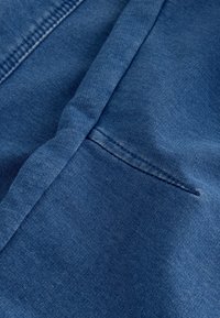 Material denim de culoare albastru închis, având o pliură și un buzunar mic, plat, cu detalii subtile de cusătură. Textură moale și suprafață uniformă.