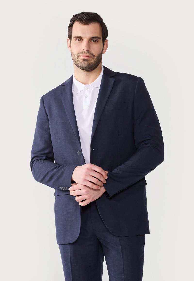 Veste de costume navy sur mesure, à bouton unique, revers crantés, deux poches avant, portée sur un polo blanc. Tissu lisse et structuré.