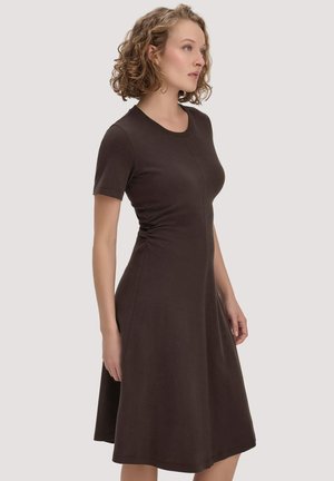 Frau mit lockigem blondem Haar, die ein eng anliegendes, kurzärmeliges, knielanges dunkelbraunes Kleid trägt und vor einem einfarbigen hellen Hintergrund steht.