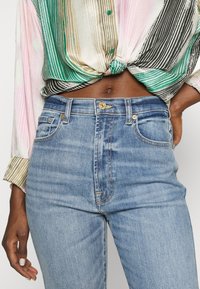 Vaaleansiniset farkut, joissa on korkea vyötärö, kultanapin kiinnitys ja hienovarainen haalistuminen. Yhdistetty monivärisen, raidallisen napitettavan paidan kanssa.