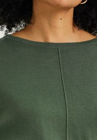 Zielony sweter dziany z szerokim dekoltem, z centralnym szwem i miękką fakturą. Materiał wydaje się lekki i lekko elastyczny.