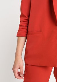 Czerwony żakiet o luźnym kroju, z pojedynczą kieszenią na piersi, podwiniętymi rękawami i gładką teksturą materiału. W parze z dopasowanymi czerwonymi spodniami.