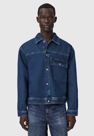 Mężczyzna w ciemnoniebieskiej dżinsowej kurtce z dużą kieszenią z klapą i dopasowanych dżinsach, stoi na tle jednolitego jasnego tła.