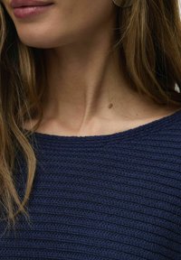 Top tricoté bleu marine avec un motif texturé en côtes et un large décolleté. Les cheveux sont de longueur moyenne et couvrent partiellement la zone du cou.