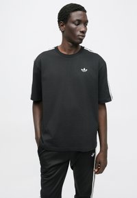 Čierne tričko s krátkymi rukávmi, okrúhlym výstrihom a bielym logom Adidas. Má biele pruhy pozdĺž ramien. Hladká zmes bavlny.