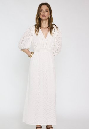 Donna che indossa un abito maxi bianco in pizzo sangallo con maniche a sbuffo, scollo a V, orecchini a cerchio dorati e braccialetti dorati coordinati.