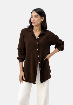 Kobieta o długich ciemnych włosach, nosząca złote kolczyki koła, brązową koszulę na guziki i białe spodnie, stojąca z jedną ręką na biodrze na białym tle.