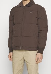 Brązowa pikowana kurtka typu puffer z kołnierzem z sztruksu, z przodu zapinana na zamek i ozdobiona pikowanym wzorem. Posiada ściągacze na mankietach oraz boczną kieszeń.