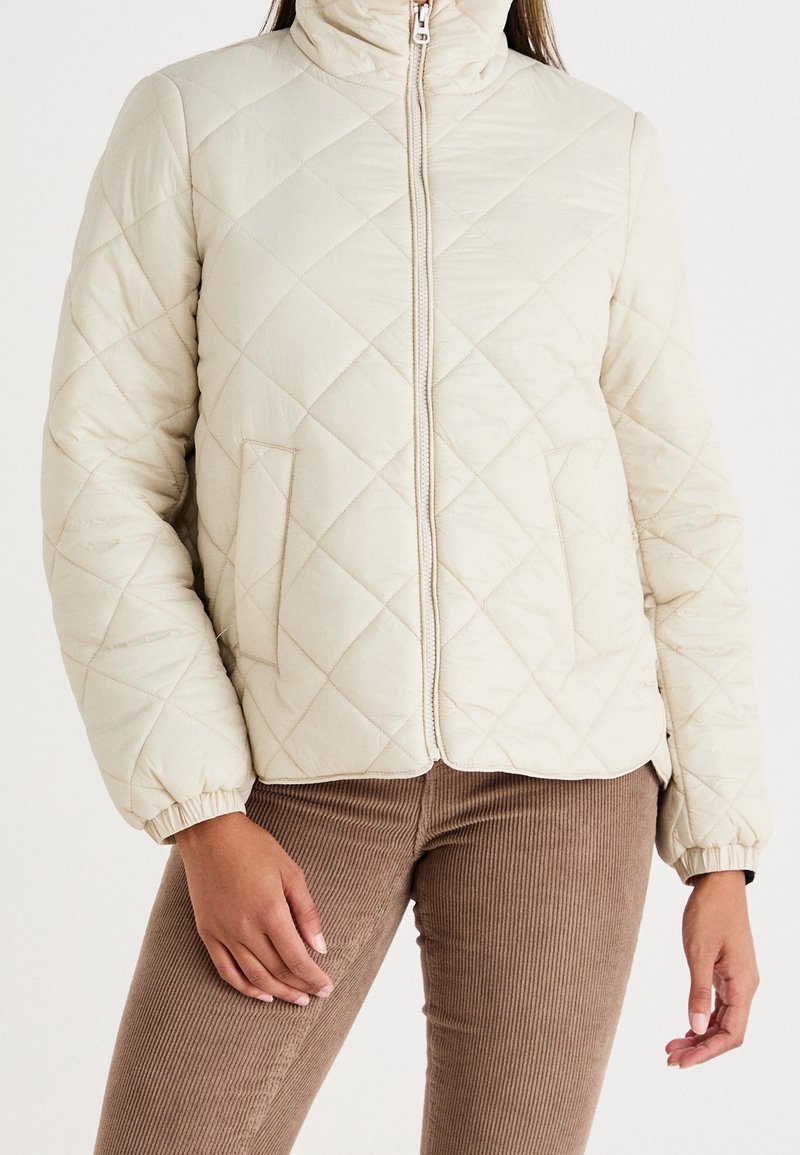 Vatteret cremefarvet dunjakke med høj krave, frontlynlås og elastiske manchetter; kombineret med brune ribstrikkede kordfløjlsbukser.