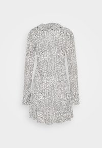 Robe blanche à manches longues avec motif de points noirs irréguliers, taille cintrée et ourlet à volants, vue de dos sur un fond uni.