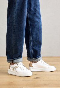 Vita sneakers med texturerade beige detaljer, sydda detaljer och en tjock vit sula, kombinerat med uppvikta mörkblå jeans.