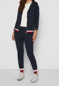 Mujer vistiendo una sudadera azul marino y joggers con puños a rayas rojas, blancas y azul marino, zapatillas blancas y una camiseta blanca debajo de la sudadera.